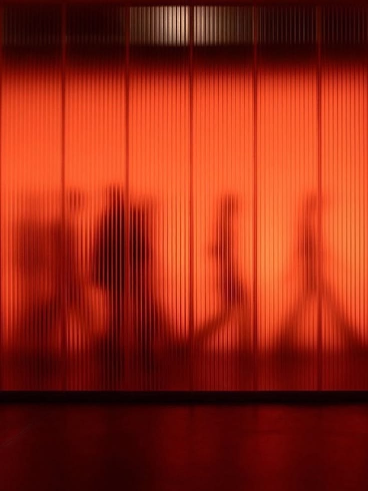 Figures gathered in a warm, amber-lit room, seen through fluted glass.